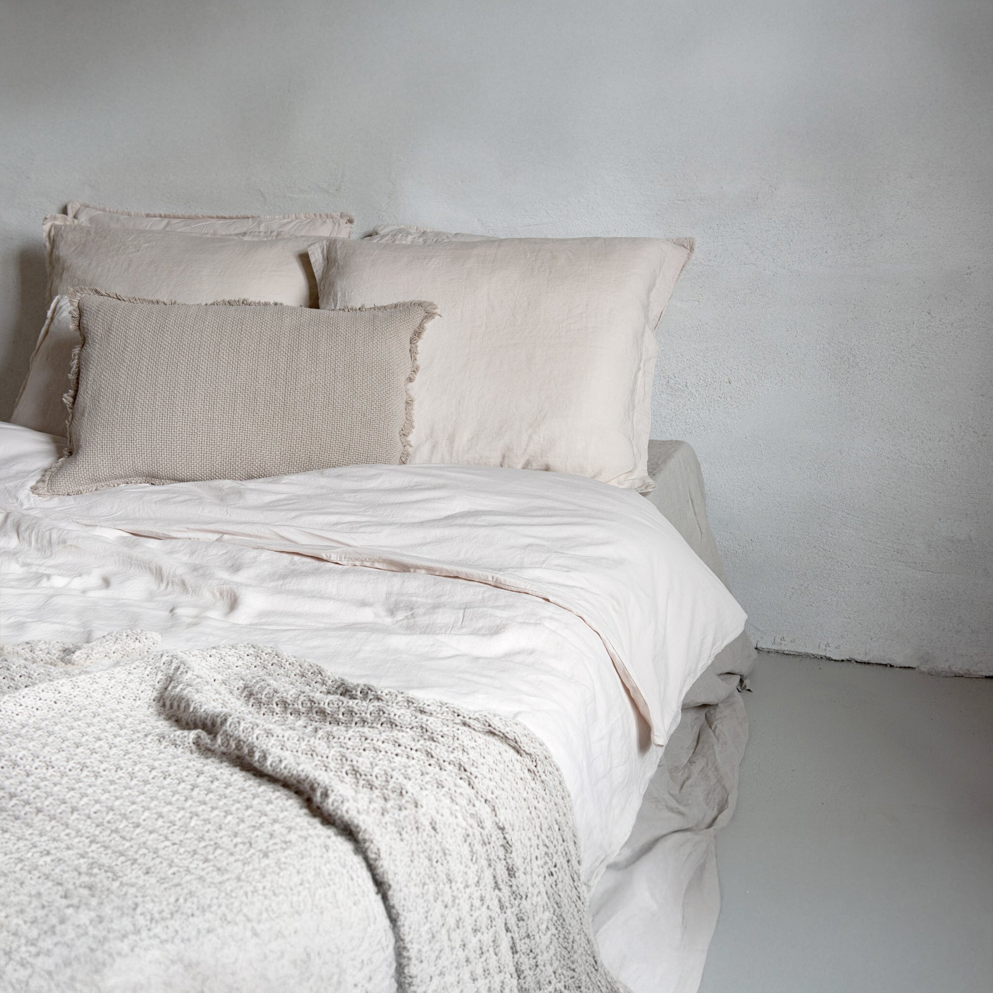 Neatly made bed with beige bedding and pillows against a plain wall.