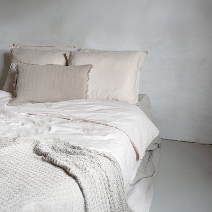 Neatly made bed with beige bedding and pillows against a plain wall.